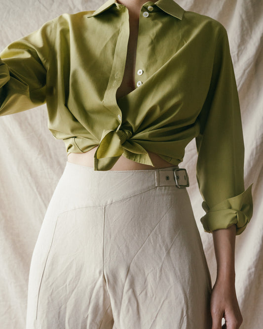 Person wearing a green vintage shirt, tied together at the front, and cream vintage trousers in front of a cream backdrop